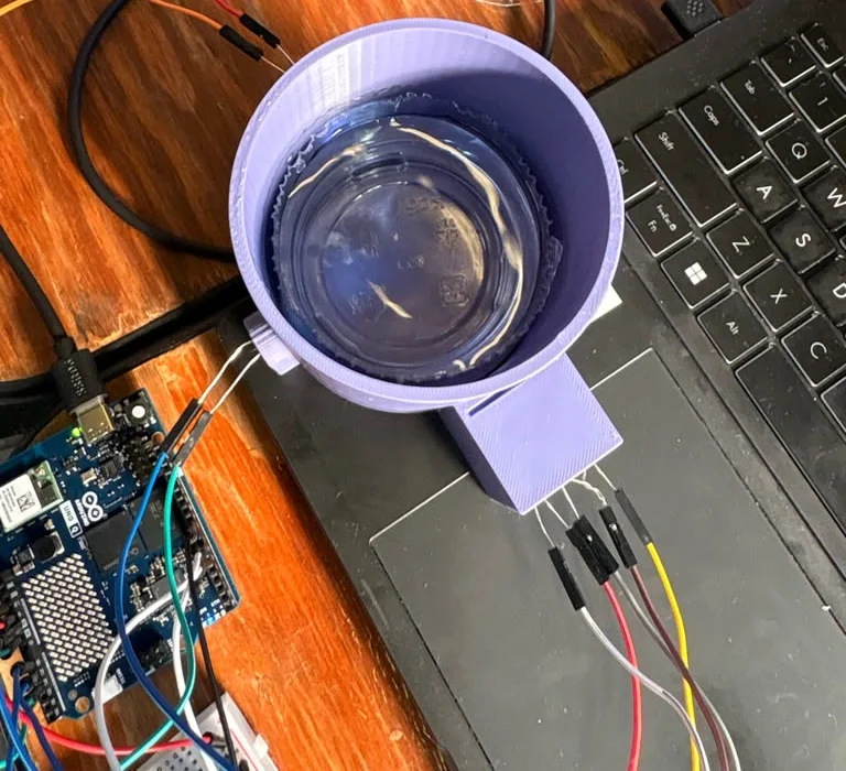 Top view with the lid off and the LEDs off: the clear plastic cup of water sitting inside the purple printed well, the LED box below it and the UNO Q at the left edge